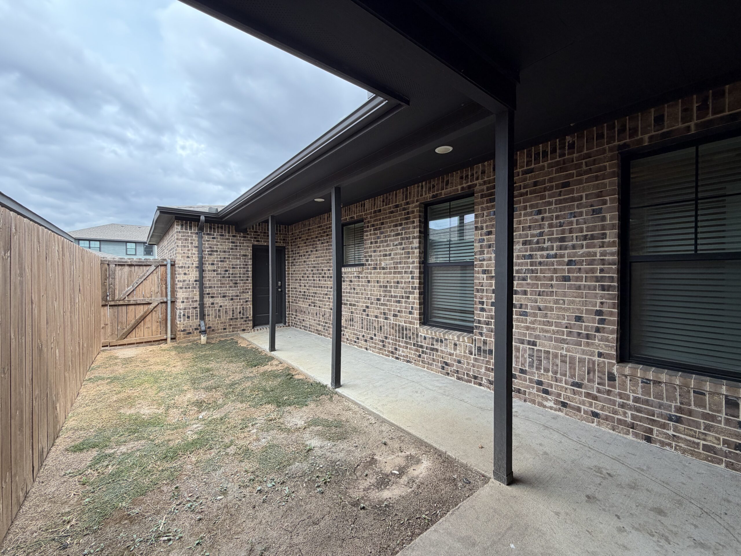 A covered walkway is visible, featuring a concrete surface and supported by black posts. The wall beside it is made of brown brick, with two windows that have dark frames and blinds. A black door is located at the end of the walkway. To the left, there is a wooden fence with a gate, and the ground is mostly bare with patches of grass. The sky is overcast, adding a muted light to the scene.