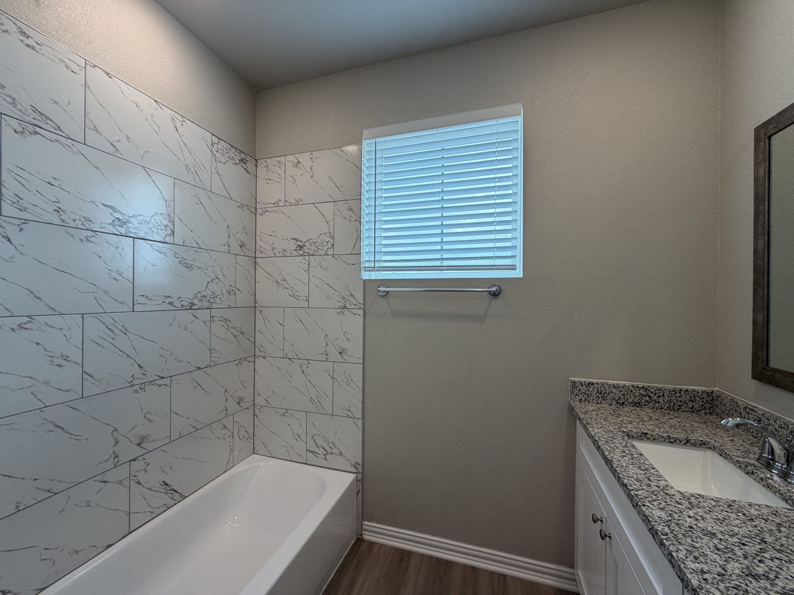 A modern bathroom features a bathtub with a white, rectangular design. The walls are adorned with large, white tiles that have a subtle marble pattern. A small window with horizontal blinds allows natural light to enter the space. To the right, there is a granite countertop with a sink and a mirror above it, enhancing the contemporary feel of the room. A towel bar is mounted on the wall near the window.