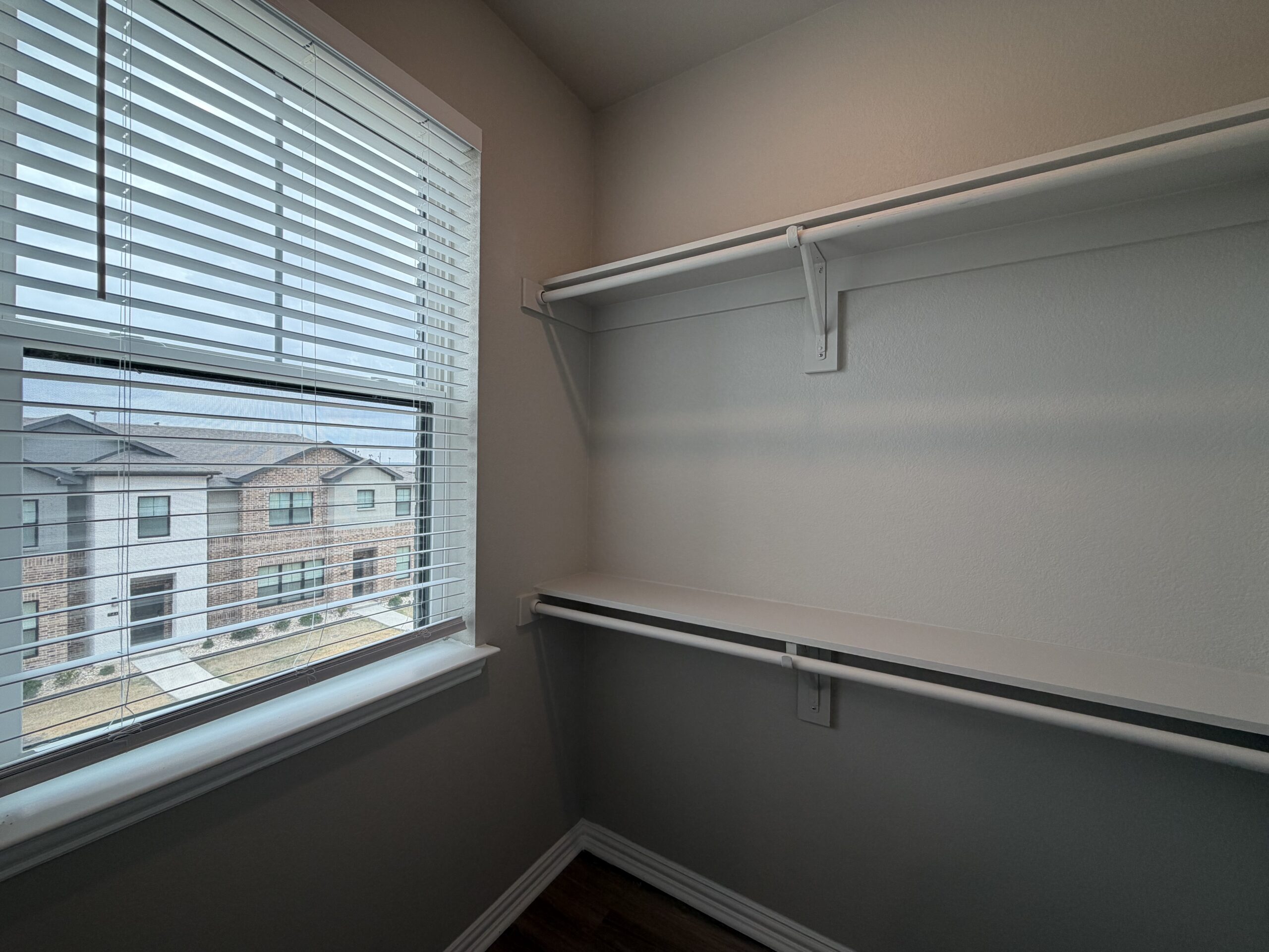 A small closet features a window with horizontal blinds, allowing natural light to enter. The walls are painted a light color, and there are two white shelves installed along the back wall. A hanging rod is positioned below the shelves, providing space for clothing or accessories. Outside the window, a view of a neighboring building is visible, showcasing a brick facade and multiple windows.