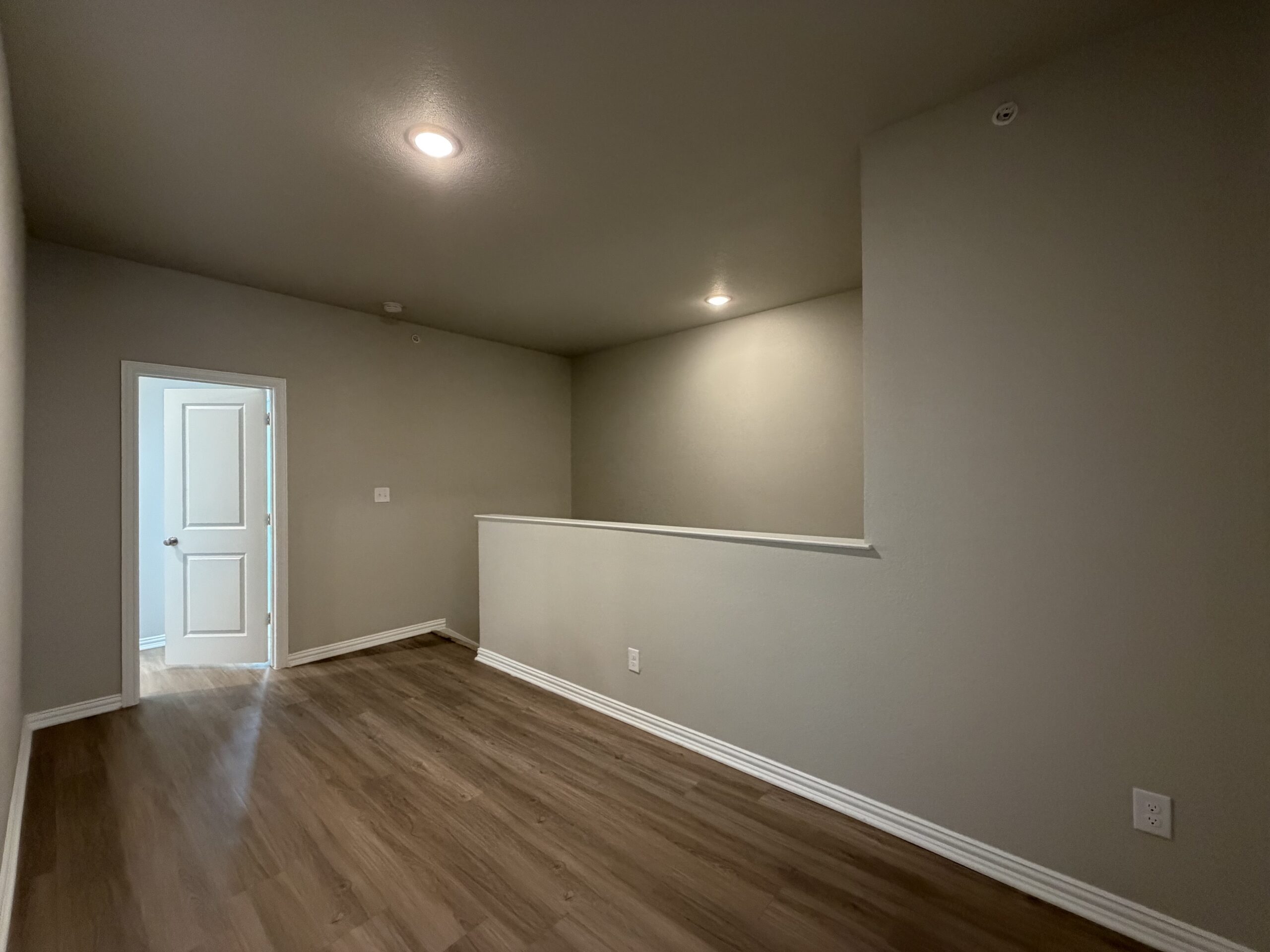 A spacious room with light gray walls and a wooden floor. The room features a doorway on the left leading to another space, and a half wall on the right that creates a separation. Recessed lighting fixtures are installed in the ceiling, providing illumination. The overall atmosphere is bright and open, with a clean and modern aesthetic.