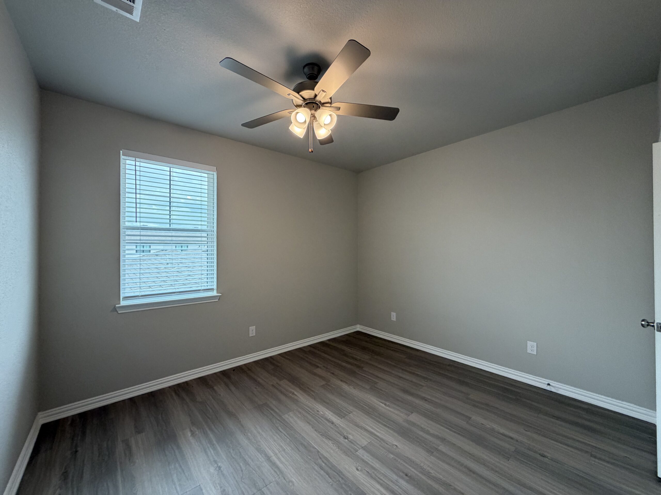 A well-lit room features light gray walls and a ceiling fan with four light bulbs. A window with white blinds allows natural light to enter, illuminating the space. The floor is covered in a wood-like laminate, and there are minimal decorations or furniture, giving the room a clean and open feel. A door is visible on the right side of the image.