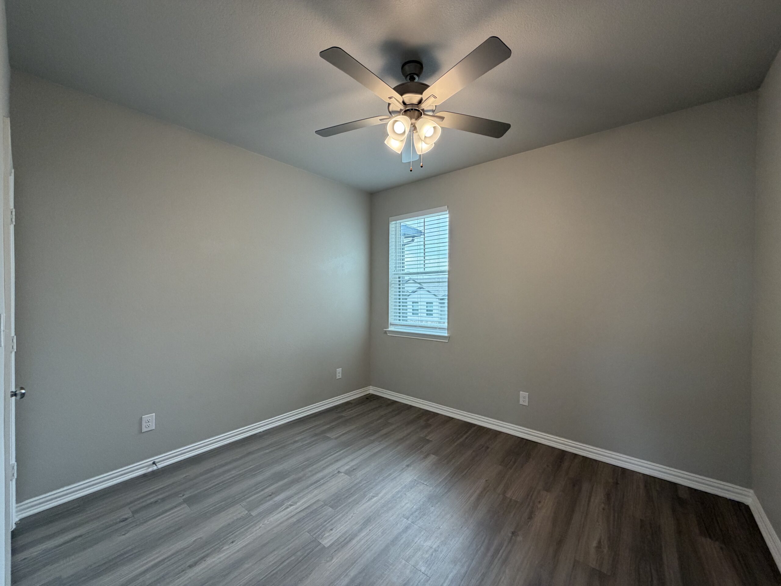 A small, empty room with light gray walls and a ceiling fan featuring three light bulbs. The floor is covered in light wood laminate. A window with blinds allows natural light to enter, revealing a view of the outside. There are electrical outlets on the walls, and a door is visible on the left side of the image. The overall atmosphere is bright and minimalistic.