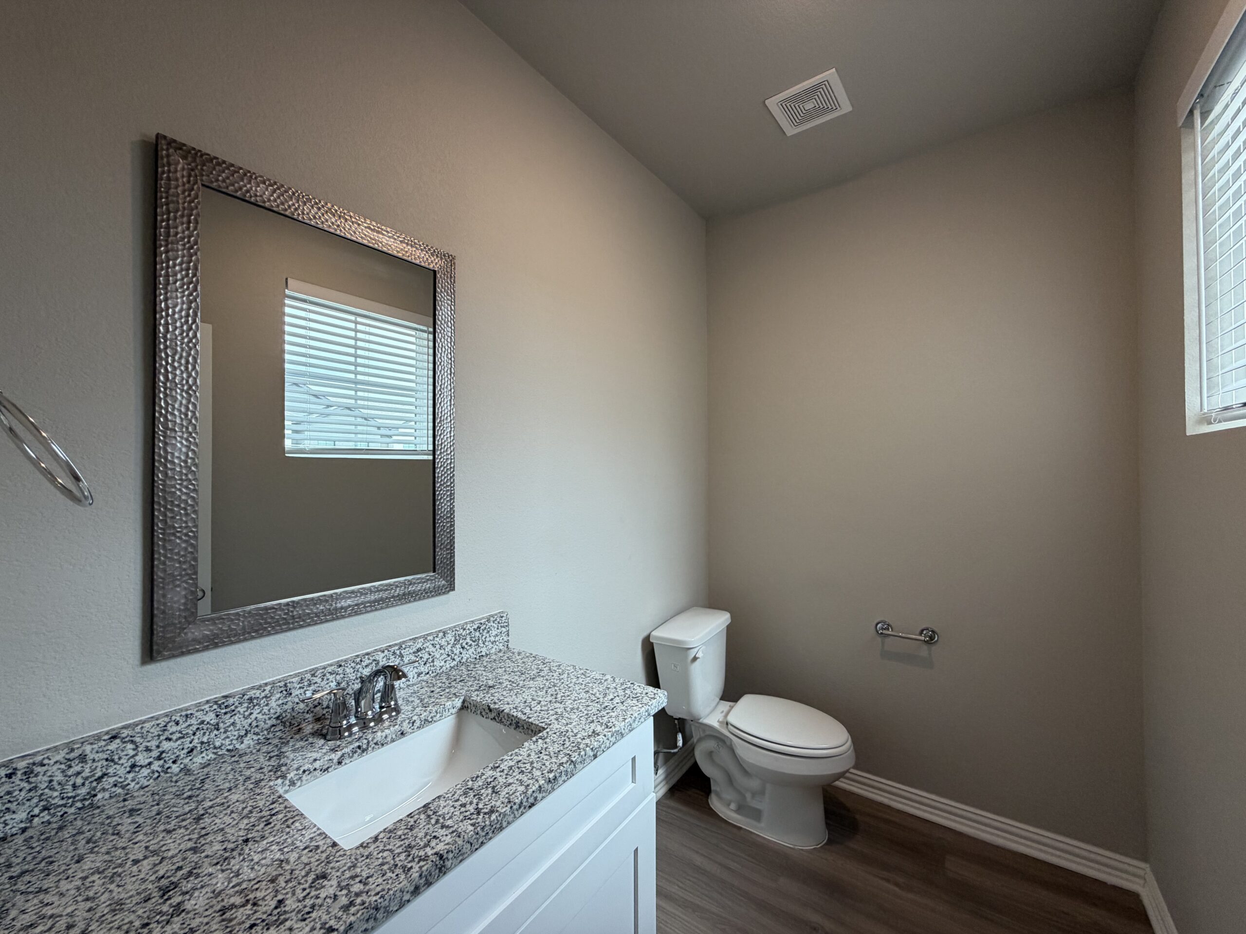 A modern bathroom features a white vanity with a granite countertop and a sleek faucet. Above the vanity is a large, framed mirror with a textured silver border. To the right, a white toilet is positioned against a light-colored wall. A small window with horizontal blinds allows natural light to enter the space, illuminating the neutral tones of the room. A towel bar is mounted on the wall near the toilet. The flooring appears to be wood-like, adding warmth to the overall design.