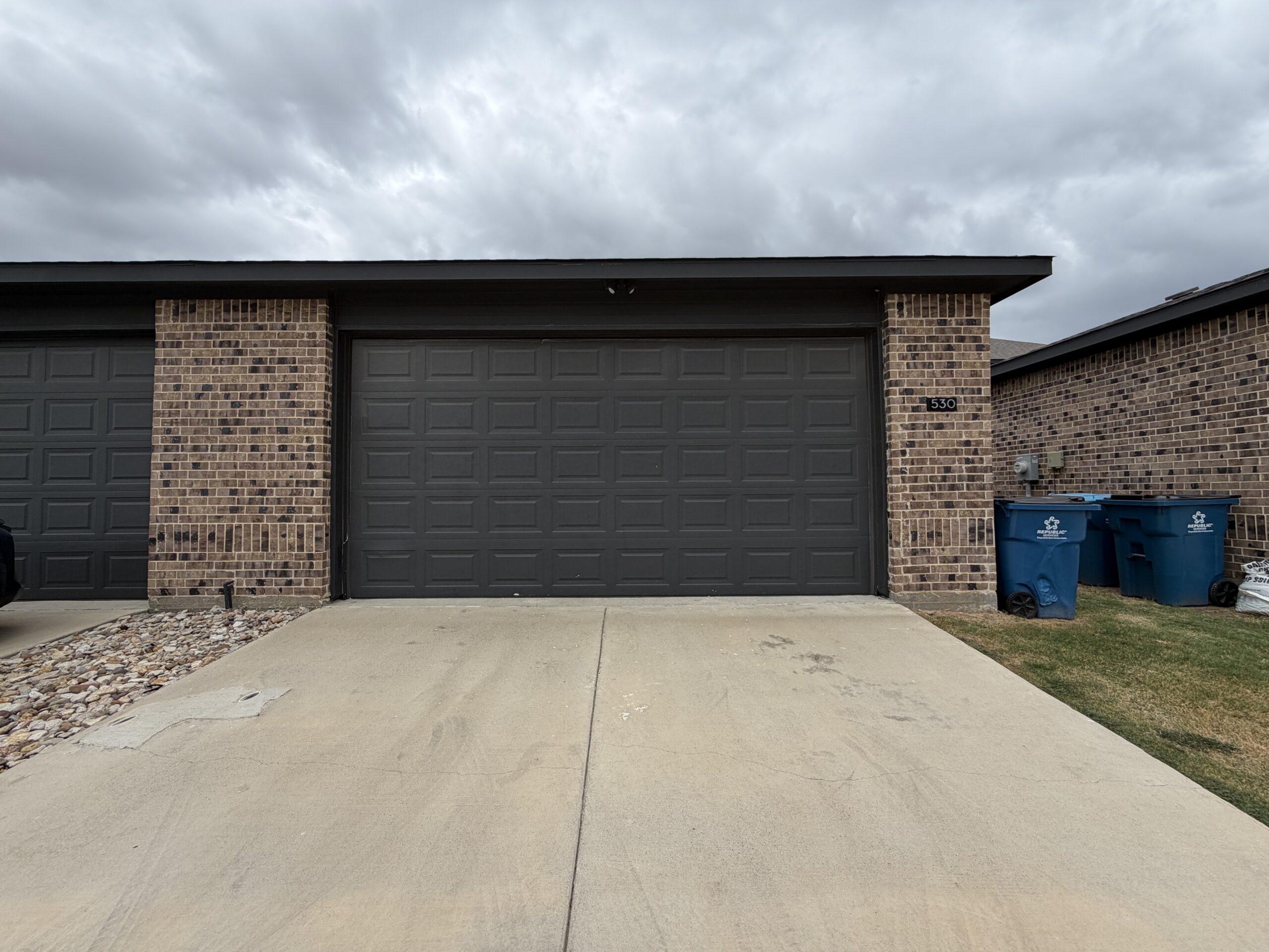 A residential garage door is prominently displayed, featuring a dark gray color with a textured panel design. To the right, there is a brick wall with the number "530" affixed to it. Next to the wall, two blue trash bins are positioned on the grass, and a small patch of gravel is visible on the left side of the driveway. The sky above is overcast, adding a muted tone to the scene.