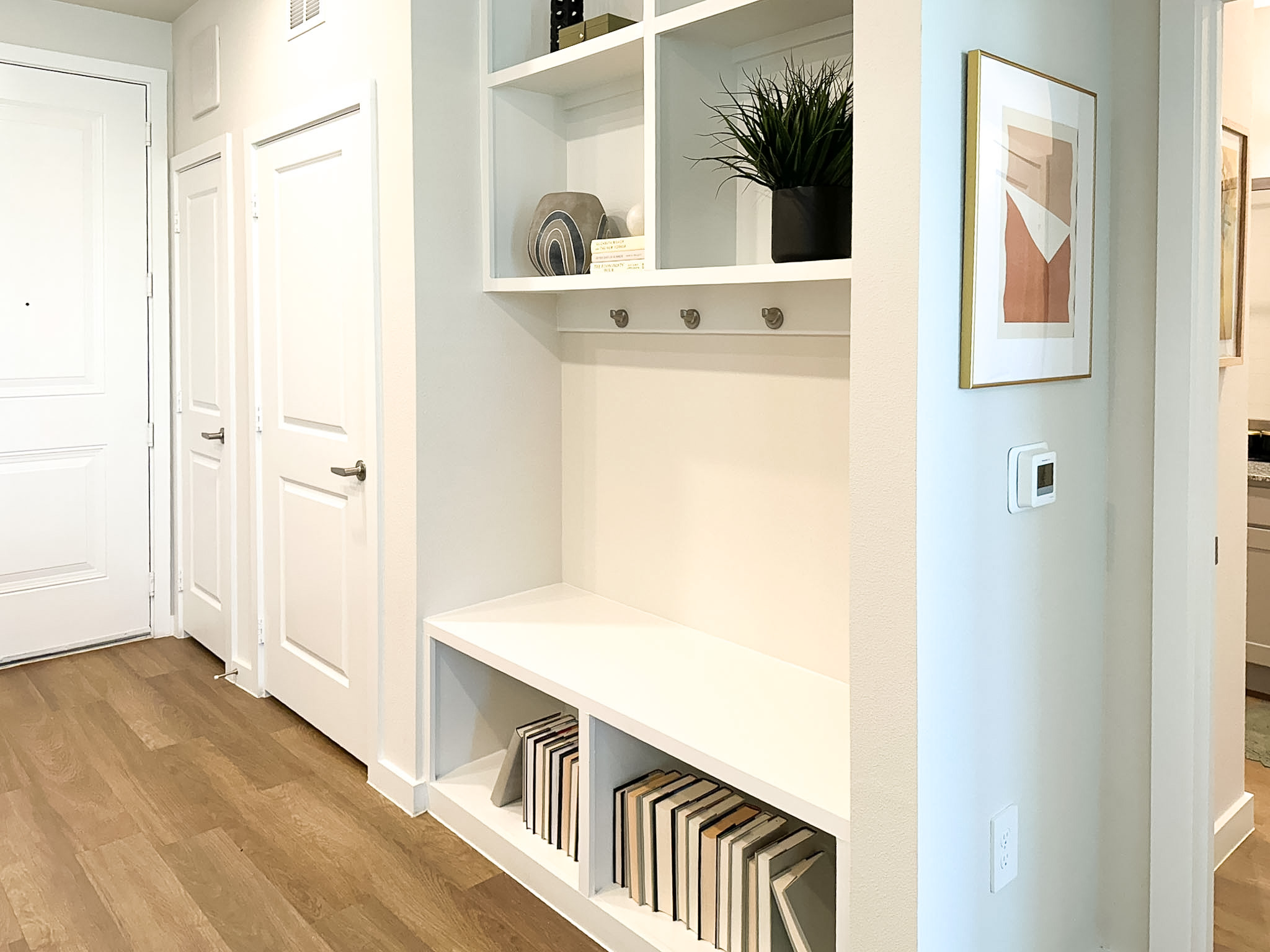 A modern entryway features a white built-in shelving unit with three hooks for hanging items. Above the hooks, there are decorative items including a small potted plant, a round decorative object, and a stack of books. The flooring is a light wood, and there are two closed doors on the left side, leading to other rooms. A framed artwork is mounted on the wall to the right, and a thermostat is visible on the wall near the shelving unit. The overall aesthetic is clean and contemporary.