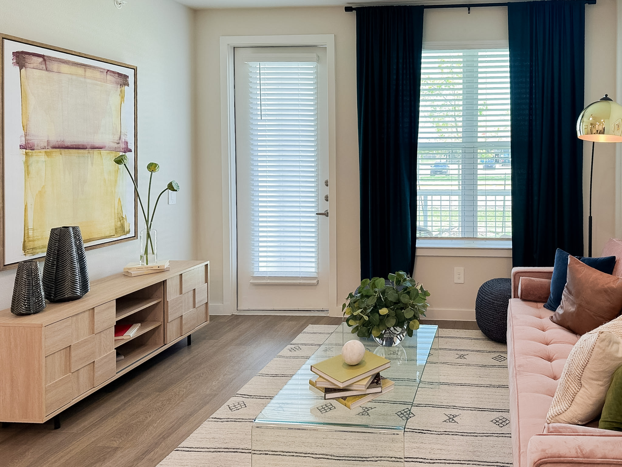 A cozy living room features a light wood media console with a textured design, holding decorative items like two black vases and a stack of books. Above it hangs a large abstract painting with yellow and purple hues. A glass coffee table sits in the center, adorned with a small white sphere and a stack of books. A green plant in a clear vase adds a touch of nature. The room has a pink sofa with various cushions, and large windows dressed with dark curtains allow natural light to filter in. A stylish floor lamp stands nearby, enhancing the inviting atmosphere.