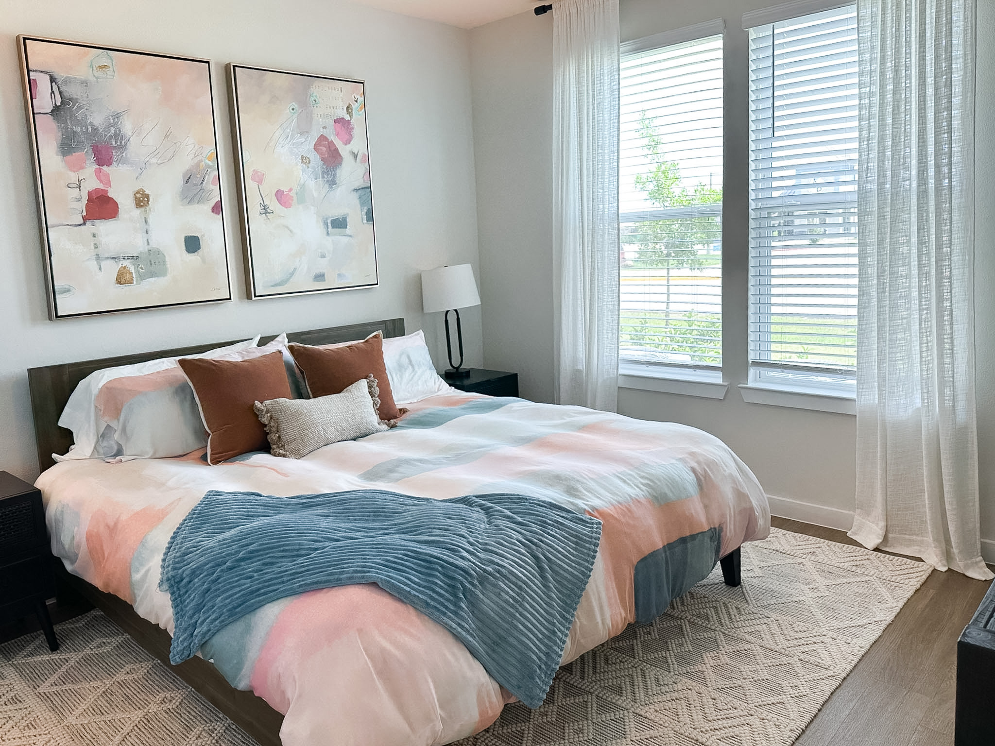 A cozy bedroom features a bed with a light, pastel-colored duvet adorned with abstract patterns. Two brown accent pillows and a textured gray pillow are placed at the head of the bed. A soft blue throw blanket is draped over the bed. On either side, there are dark nightstands, one with a modern lamp. Large windows with white blinds allow natural light to fill the room, and sheer curtains add a soft touch. Two abstract paintings hang above the bed, enhancing the room's aesthetic. A light-colored area rug is positioned on the wooden floor.