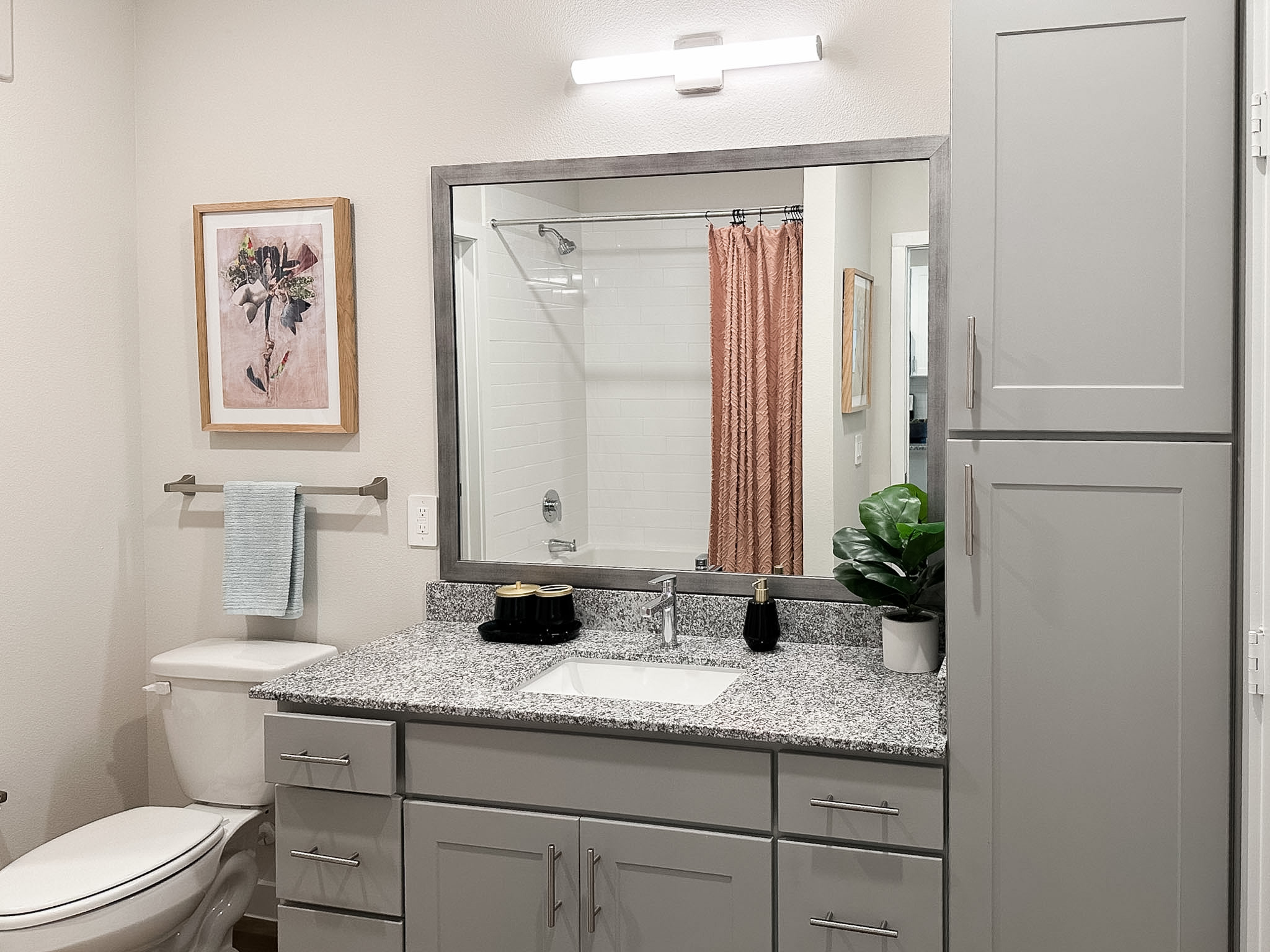 A modern bathroom features a large mirror with a gray frame above a granite countertop. The countertop holds a small black tray with decorative items and a soap dispenser. A white sink is integrated into the countertop. To the left, a white toilet is positioned next to a towel rack with a light blue towel. A peach-colored shower curtain hangs in the background, partially obscuring a white tiled shower. A framed artwork with floral elements is mounted on the wall, adding a touch of color. A small potted plant sits on the countertop, enhancing the decor.