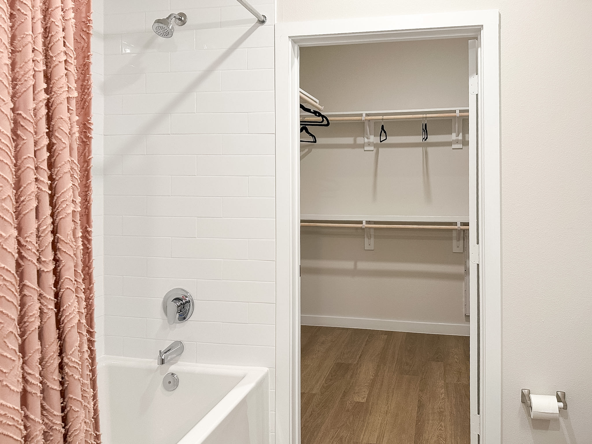A bathroom scene features a pink textured shower curtain on the left, partially covering a white tiled wall. In the foreground, a bathtub faucet is visible. To the right, an open closet reveals a simple design with two wooden shelves and a couple of black hangers. The floor is finished with light wood, and the walls are painted a neutral color.