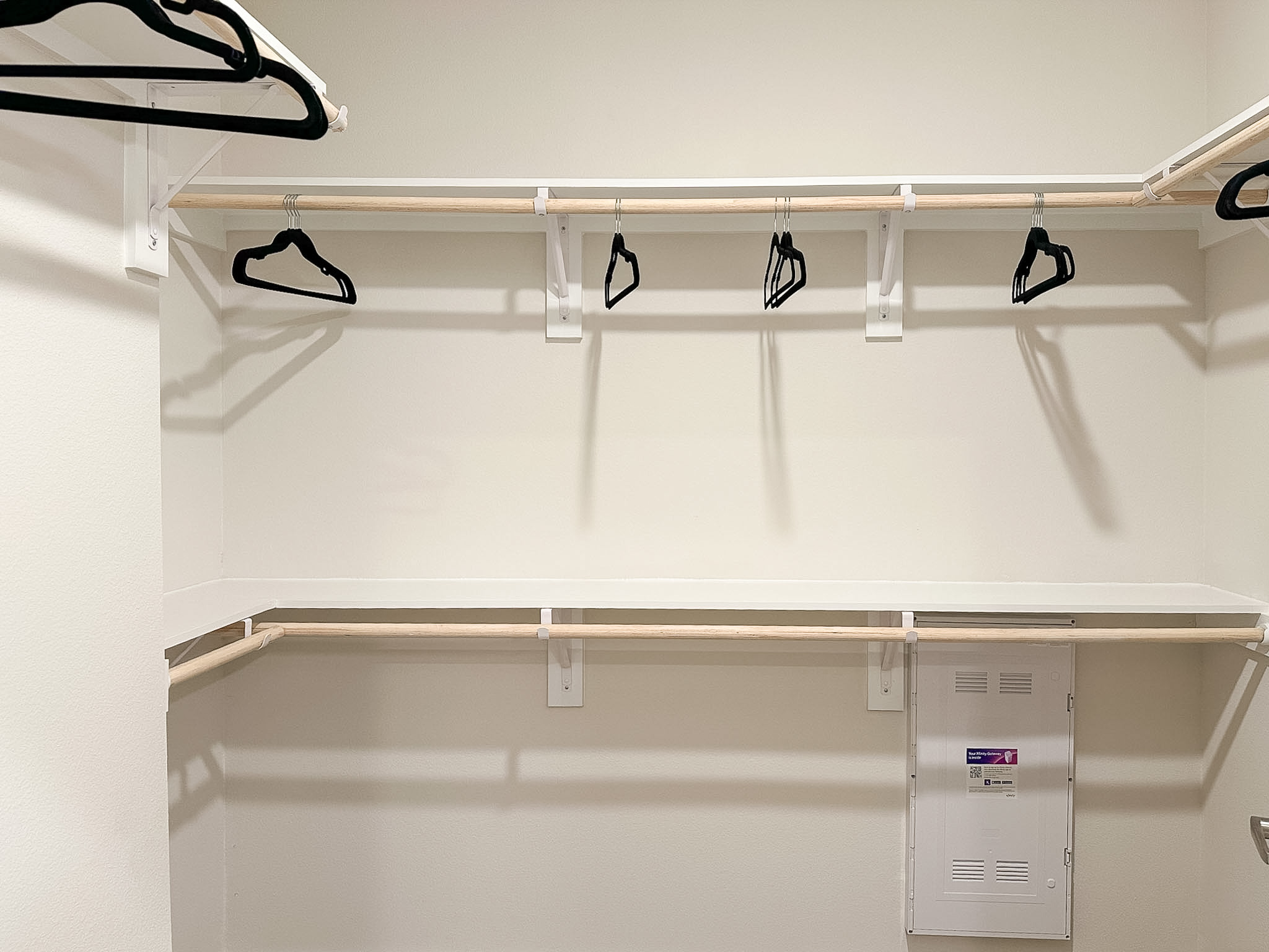 A spacious closet features a light-colored wall and a wooden hanging rod supported by white brackets. Several black hangers are suspended from the rod. Below, a white shelf runs along the back wall, and a small panel is visible on the lower right side, likely for access to utilities. The overall design is simple and functional, with a clean and organized appearance.