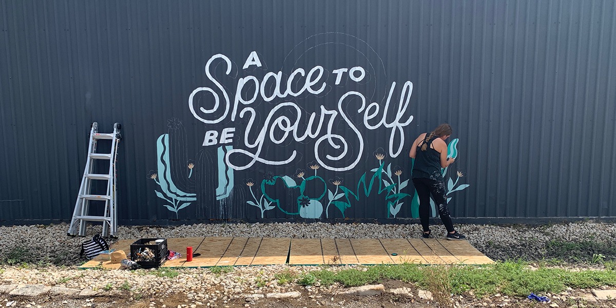 woman painting "a space to be yourself" mural on side of warehouse